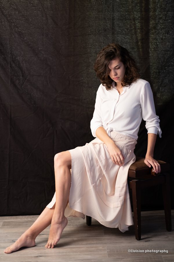 Seated studio portrait in a white shirt and pale linen skirt against a dark backdrop