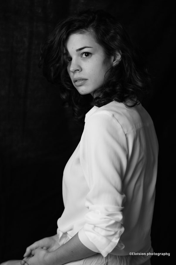 Seated studio portrait in a white shirt and pale linen skirt against a dark backdrop