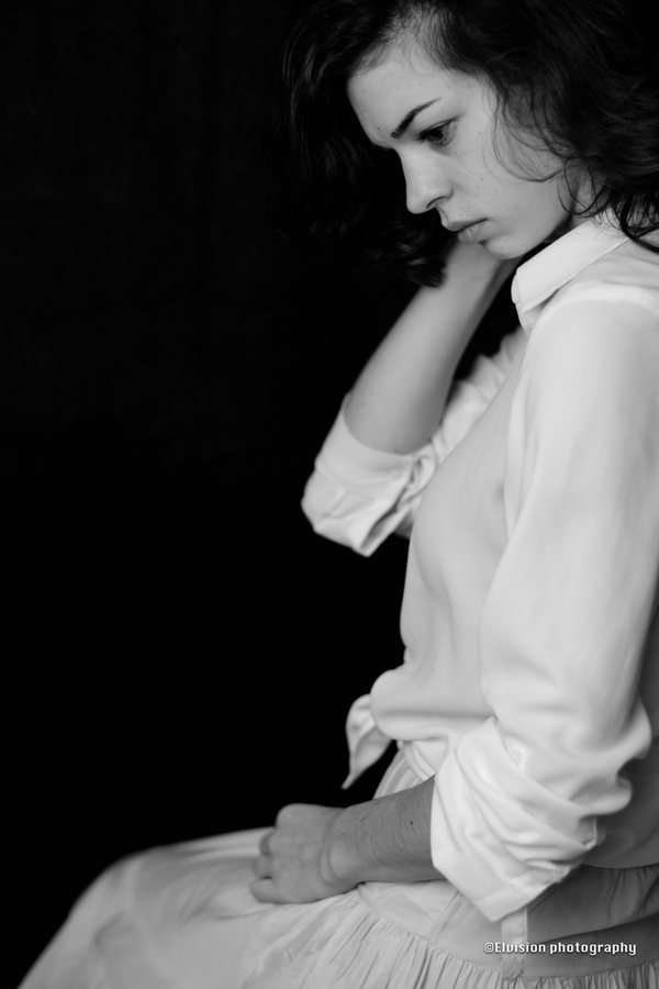 Seated studio portrait in a white shirt and pale linen skirt against a dark backdrop