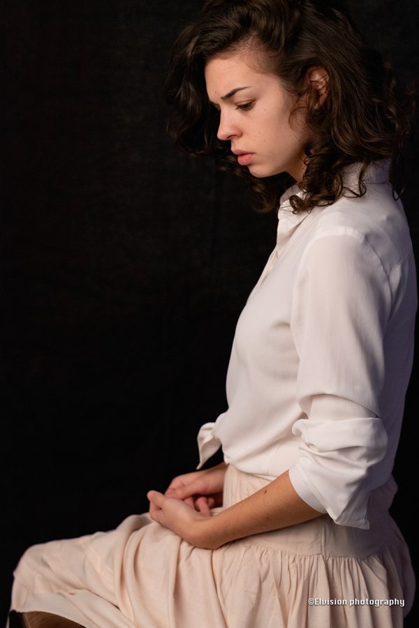 Seated studio portrait in a white shirt and pale linen skirt against a dark backdrop