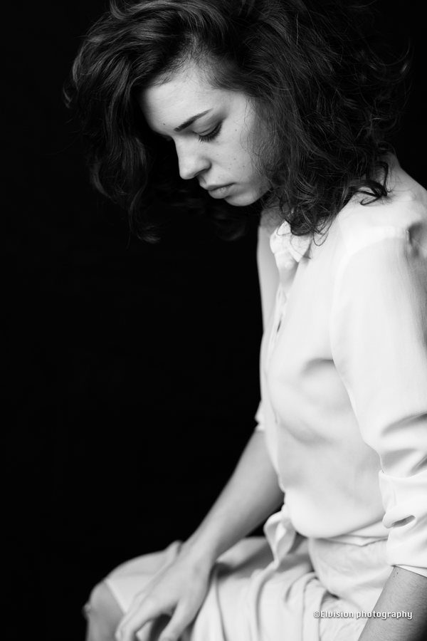 Seated studio portrait in a white shirt and pale linen skirt against a dark backdrop