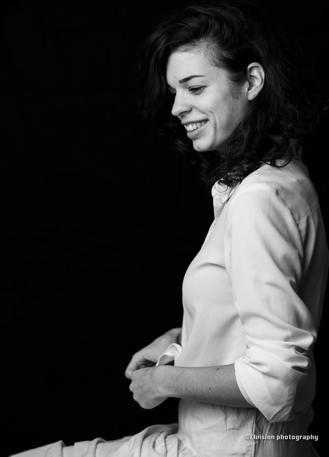 Seated studio portrait in a white shirt and pale linen skirt against a dark backdrop