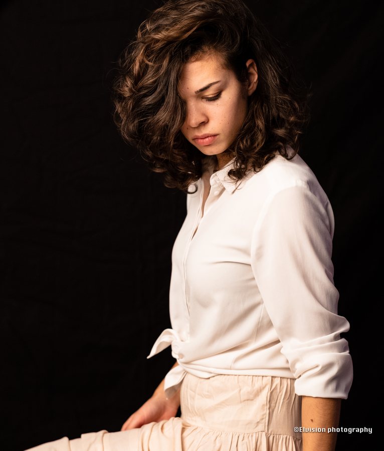 Seated studio portrait in a white shirt and pale linen skirt against a dark backdrop