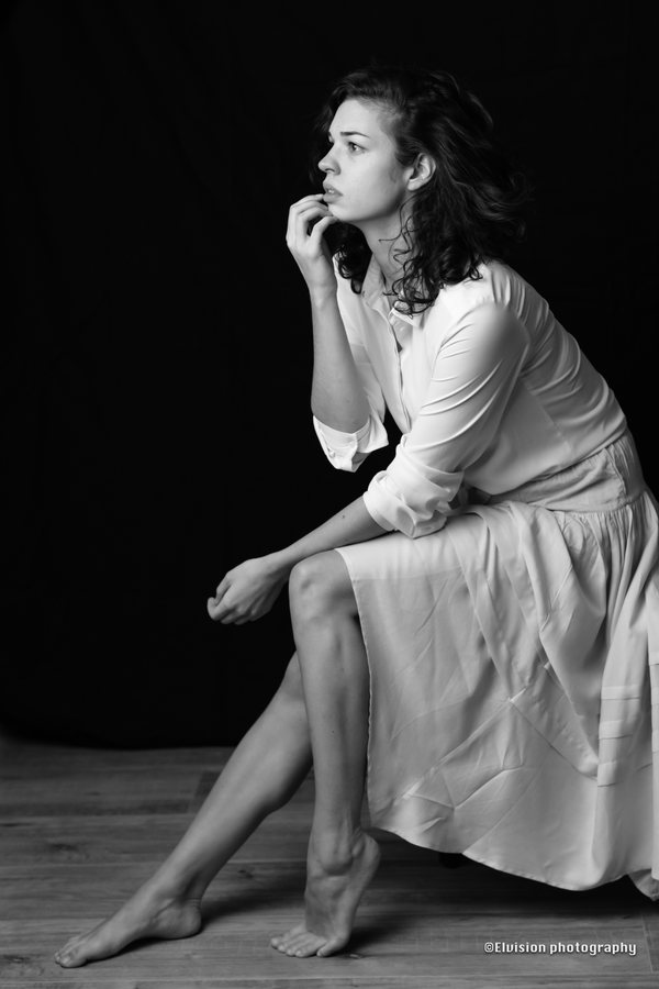 Seated studio portrait in a white shirt and pale linen skirt against a dark backdrop