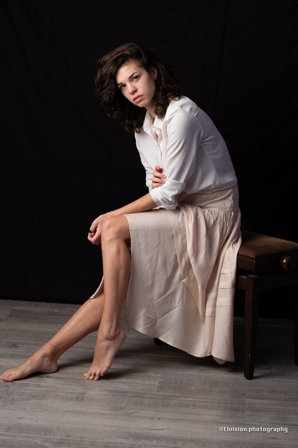 Seated studio portrait in a white shirt and pale linen skirt against a dark backdrop